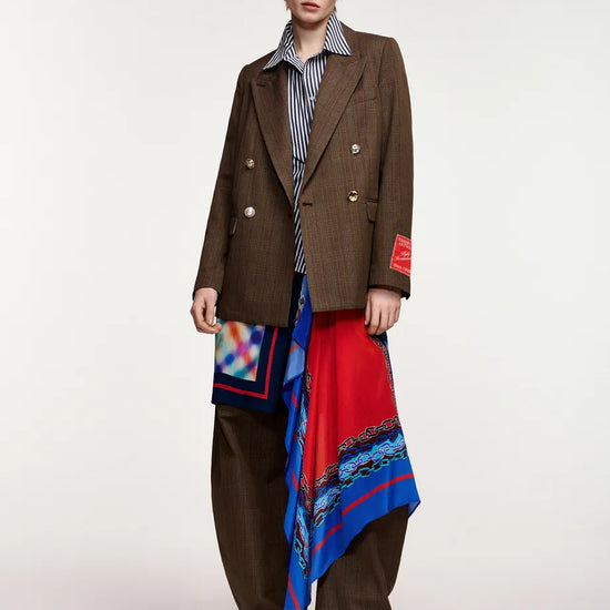 A model stands against a light gray studio background wearing a brown double-breasted pinstripe blazer, matching brown trousers, a blue-and-white striped shirt, and a long red scarf with blue and multicolor patterns draped over the lower body.