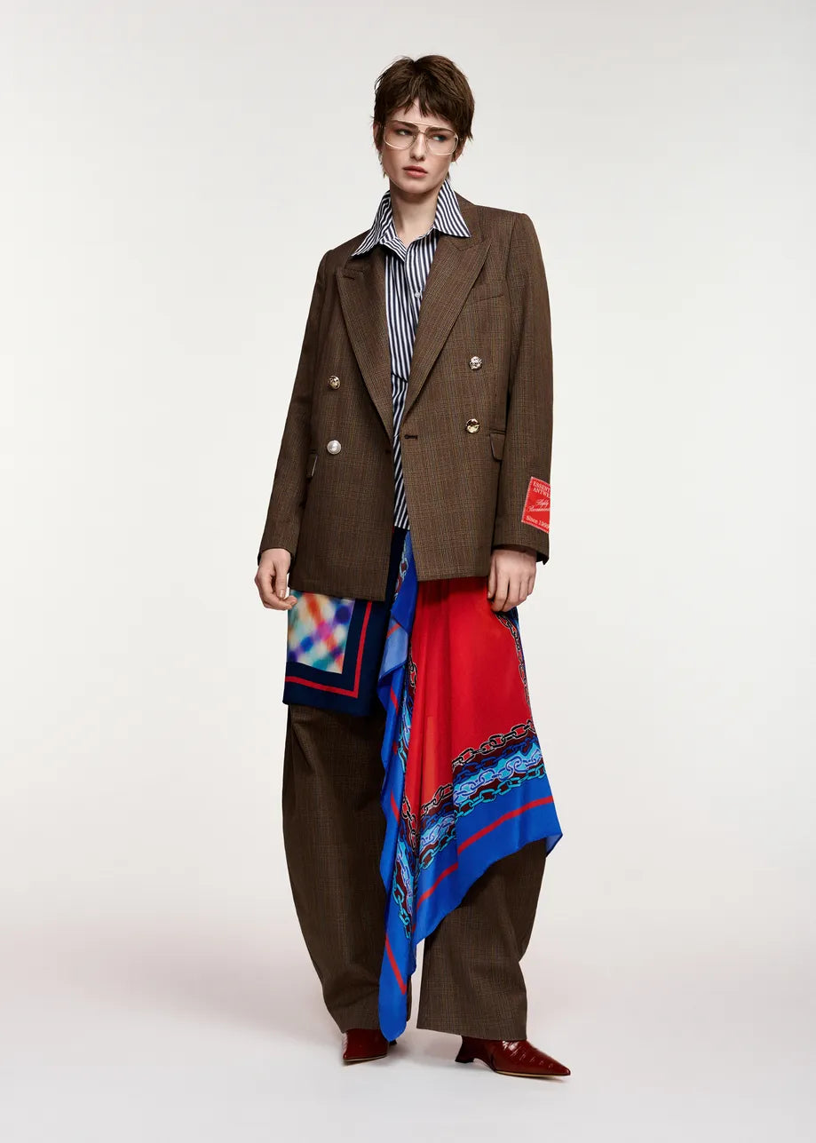 A model stands against a light gray studio background wearing a brown double-breasted pinstripe blazer, matching brown trousers, a blue-and-white striped shirt, and a long red scarf with blue and multicolor patterns draped over the lower body.