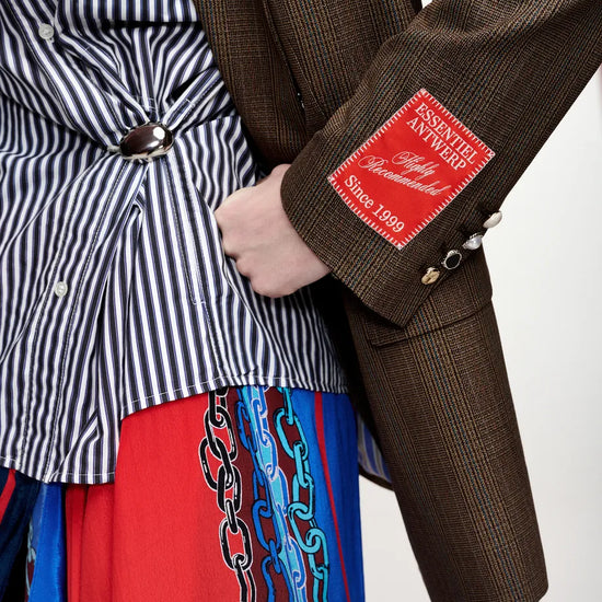 Close-up of a person wearing a brown textured blazer with a red patch on the sleeve, layered over a black-and-white striped shirt and a red skirt with blue chain embroidery.