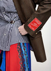 Close-up of a person wearing a brown textured blazer with a red patch on the sleeve, layered over a black-and-white striped shirt and a red skirt with blue chain embroidery.