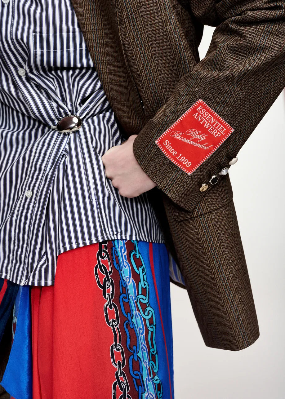 Close-up of a person wearing a brown textured blazer with a red patch on the sleeve, layered over a black-and-white striped shirt and a red skirt with blue chain embroidery.