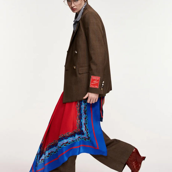 A model walks in profile wearing a brown double-breasted blazer, matching brown trousers, a striped shirt, and a long red and blue patterned scarf draped over one leg, paired with brown boots, against a plain light gray studio background.