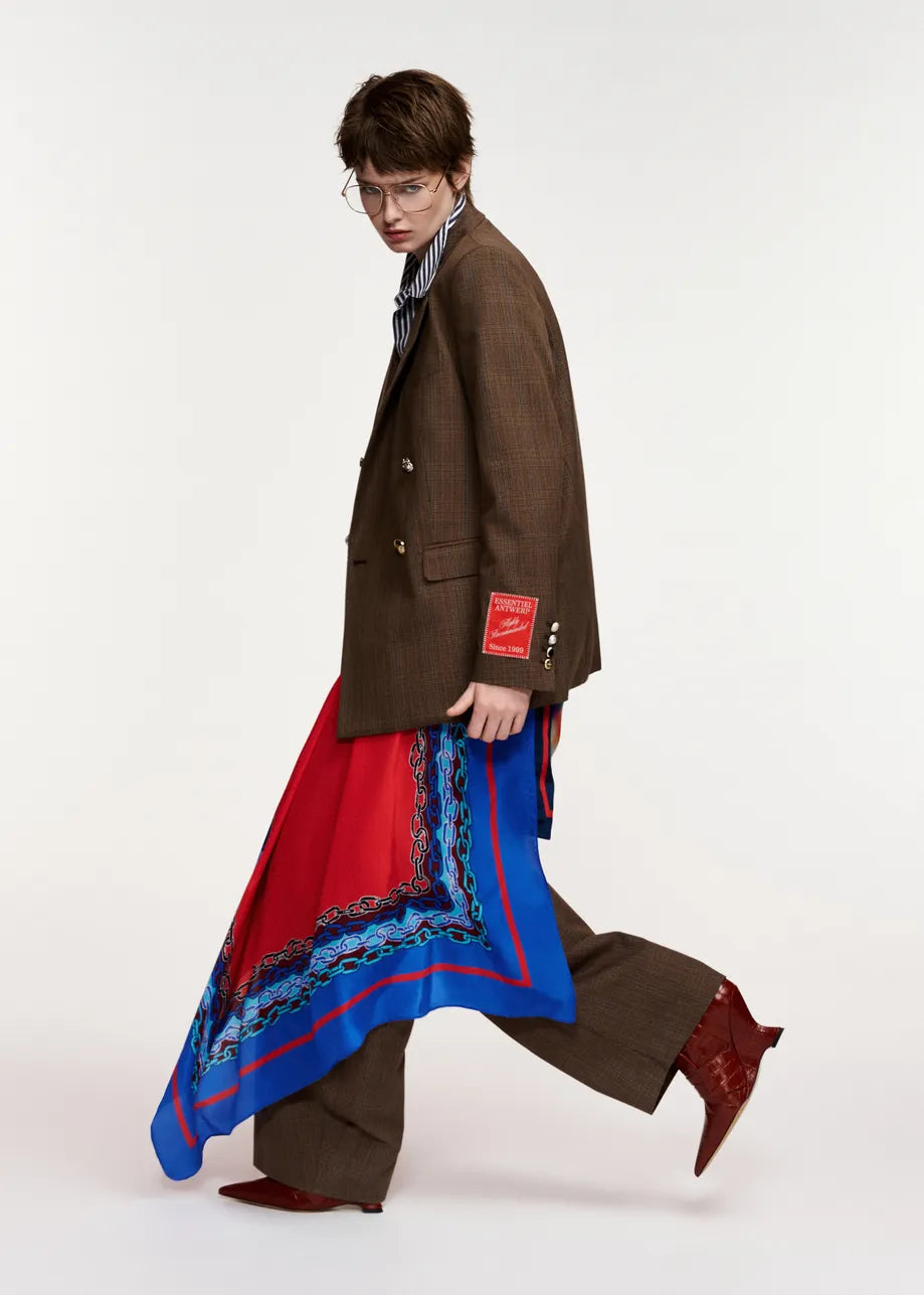 A model walks in profile wearing a brown double-breasted blazer, matching brown trousers, a striped shirt, and a long red and blue patterned scarf draped over one leg, paired with brown boots, against a plain light gray studio background.