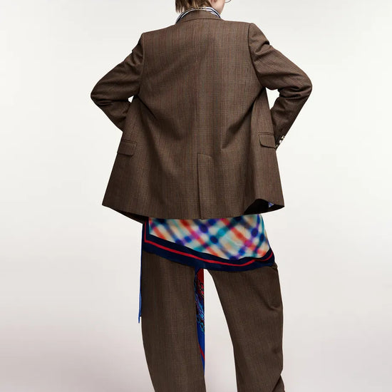 A model shown from the back wearing an oversized brown pinstripe blazer, matching wide-leg trousers, a colorful plaid shirt, and red pointed shoes against a plain white studio background.
