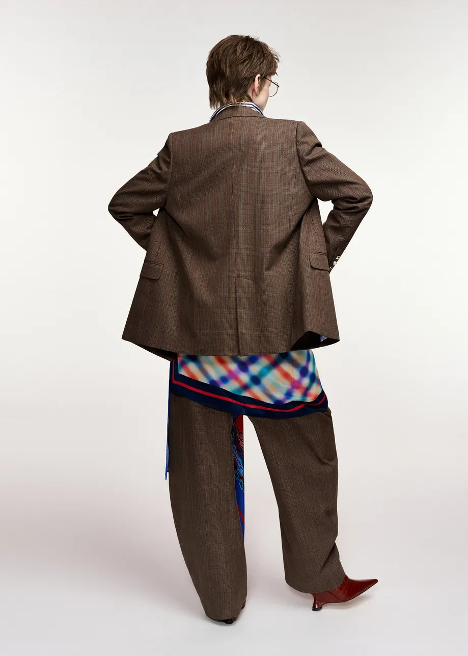 A model shown from the back wearing an oversized brown pinstripe blazer, matching wide-leg trousers, a colorful plaid shirt, and red pointed shoes against a plain white studio background.