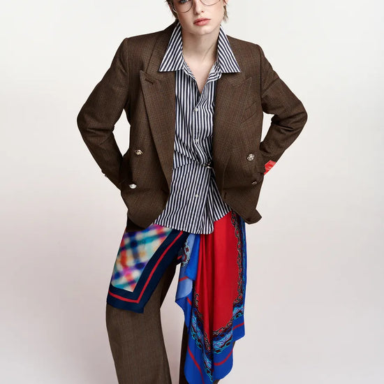 A model wearing a brown double-breasted blazer, a blue and white striped button-down shirt, wide-leg brown trousers, and a vibrant red, blue, and black patterned silk scarf draped over the lap and waist, set against a plain light gray studio background.