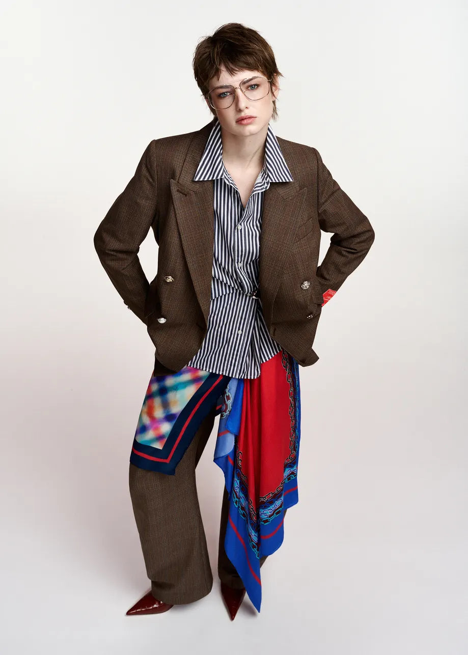A model wearing a brown double-breasted blazer, a blue and white striped button-down shirt, wide-leg brown trousers, and a vibrant red, blue, and black patterned silk scarf draped over the lap and waist, set against a plain light gray studio background.