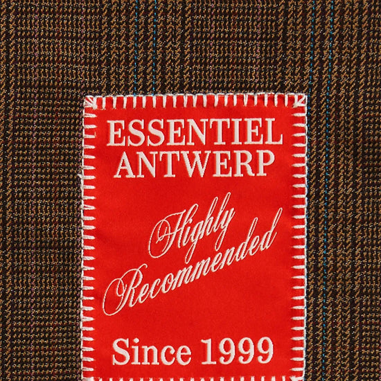 A close-up of a brown and black plaid fabric with a prominent red rectangular patch reading 'ESSENTIEL ANTWERP Highly Recommended Since 1999' in white text.