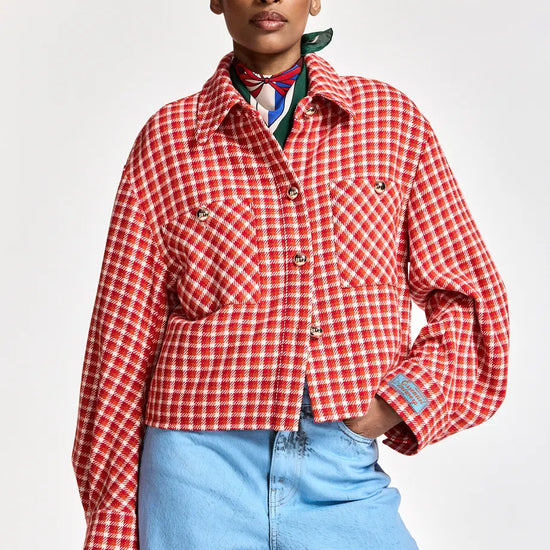 A model wearing a red and white checkered cropped jacket with front pockets, styled with a colorful neck scarf and light blue jeans against a plain light gray background.