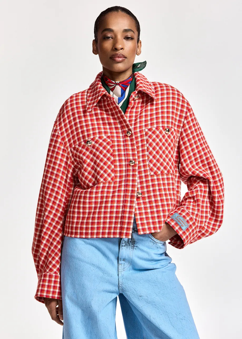 A model wearing a red and white checkered cropped jacket with front pockets, styled with a colorful neck scarf and light blue jeans against a plain light gray background.