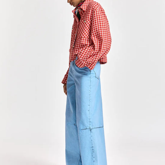 A model is shown from the side wearing a red and white gingham button-up shirt, light blue wide-leg denim jeans, and red shoes, set against a plain light gray studio background.