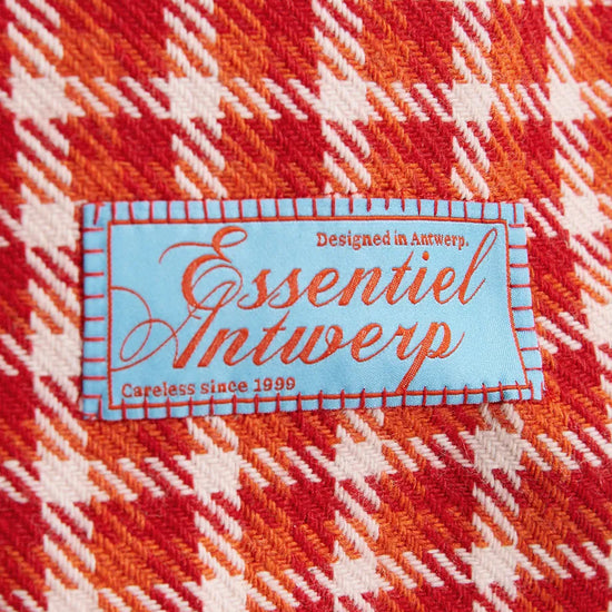 A close-up of a red, orange, and white plaid fabric with a central light blue sewn label reading 'Essentiel Antwerp'.
