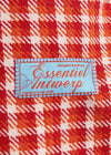 A close-up of a red, orange, and white plaid fabric with a central light blue sewn label reading 'Essentiel Antwerp'.