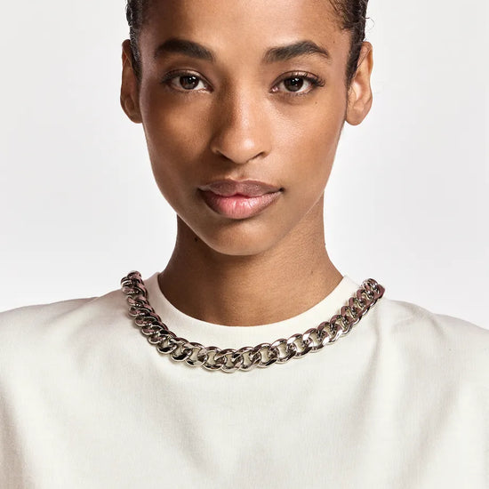 A close-up portrait of a model wearing a chunky silver-toned chain necklace over a white crewneck top, set against a plain light gray background.