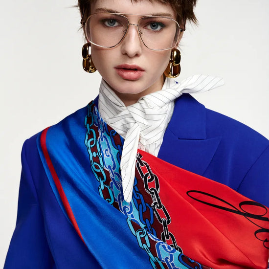 A model with short brown hair and glasses wearing a vibrant blue blazer, a white striped necktie, and a bright red and blue patterned scarf with a chain-link design, set against a plain light gray background.