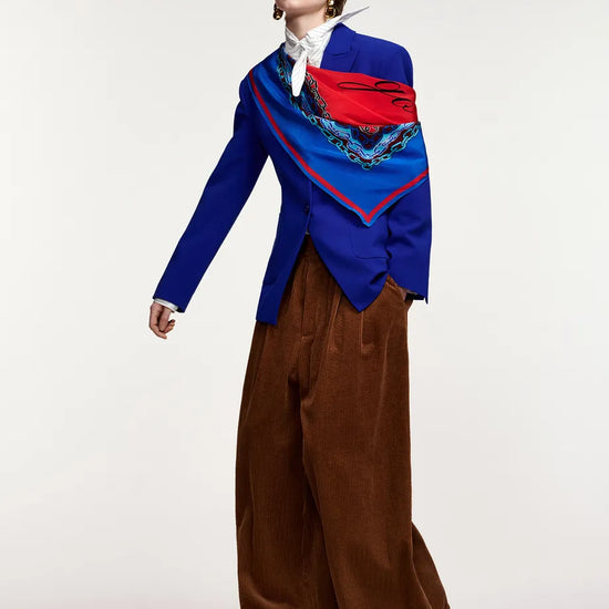 A model wearing a vibrant blue blazer with a red and blue patterned scarf draped over the shoulders, paired with wide-leg brown trousers, a white shirt, and red shoes, photographed against a plain light gray studio background.
