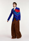 A model wearing a vibrant blue blazer with a red and blue patterned scarf draped over the shoulders, paired with wide-leg brown trousers, a white shirt, and red shoes, photographed against a plain light gray studio background.