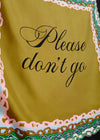 A close-up of a yellow-green fabric item, likely a scarf or sash, featuring the phrase 'Please don't go' in black cursive script. The fabric is finished with a decorative border in pink, green, and gold.