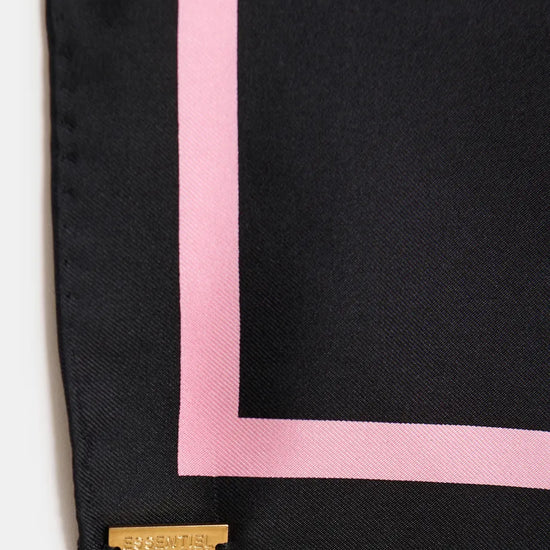 A close-up of a black silk scarf with a pink border and a gold-tone metal brand plaque at the corner.