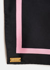 A close-up of a black silk scarf with a pink border and a gold-tone metal brand plaque at the corner.