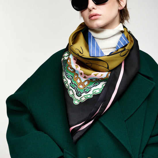 A model wearing a dark green wool coat, a white turtleneck, and a patterned silk scarf in olive green, black, blue, and white, styled with oversized black sunglasses against a light gray background.