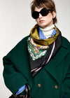 A model wearing a dark green wool coat, a white turtleneck, and a patterned silk scarf in olive green, black, blue, and white, styled with oversized black sunglasses against a light gray background.