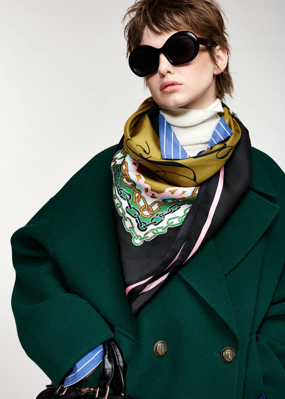 A model wearing a dark green wool coat, a white turtleneck, and a patterned silk scarf in olive green, black, blue, and white, styled with oversized black sunglasses against a light gray background.