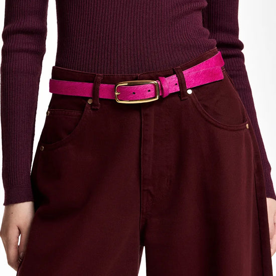 A close-up of a person wearing a bright pink belt with a gold-tone buckle over a burgundy ribbed long-sleeve top and matching wide-leg trousers.
