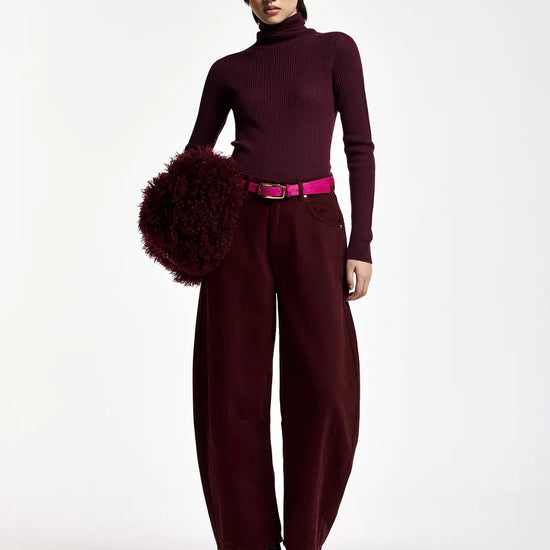 A model stands against a plain white studio background wearing a dark burgundy turtleneck sweater, matching wide-leg trousers, a bright pink belt, black pointed boots, and holding a large dark burgundy fuzzy pom-pom accessory.