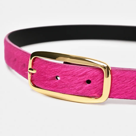 A close-up of a belt with a textured hot pink strap, a black interior lining, and a polished gold-tone metal buckle.