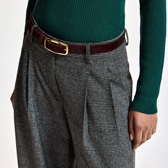 A close-up of a person wearing gray and black plaid wide-leg trousers with pleats, a dark brown leather belt with a gold buckle, and a dark green ribbed knit sweater.
