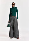 A model wearing a dark green ribbed turtleneck sweater, wide-leg gray pleated trousers with a brown belt, sunglasses, and holding a green handbag against a plain white studio background.