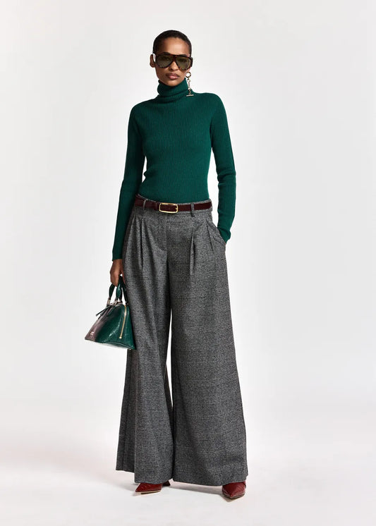 A model wearing a dark green ribbed turtleneck sweater, wide-leg gray pleated trousers with a brown belt, sunglasses, and holding a green handbag against a plain white studio background.