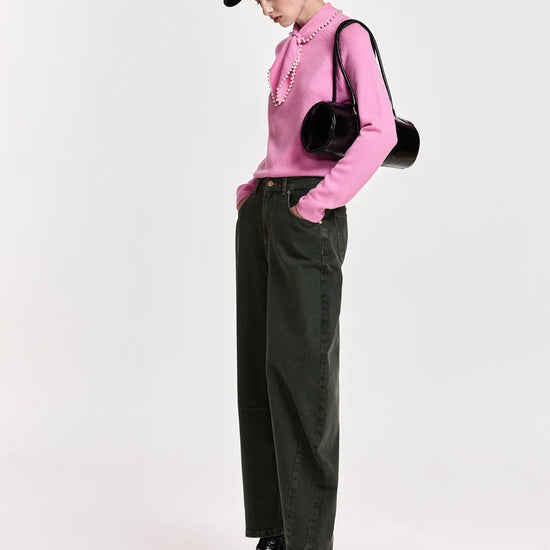 A model wearing a pink long-sleeve knit top with a ruffled collar, dark green wide-leg jeans, black patent leather ankle boots, a black baseball cap, and a black round shoulder bag, standing against a light gray studio background.