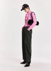 A model wearing a pink long-sleeve knit top with a ruffled collar, dark green wide-leg jeans, black patent leather ankle boots, a black baseball cap, and a black round shoulder bag, standing against a light gray studio background.