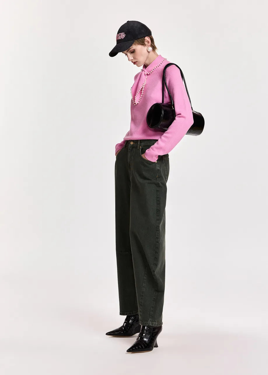 A model wearing a pink long-sleeve knit top with a ruffled collar, dark green wide-leg jeans, black patent leather ankle boots, a black baseball cap, and a black round shoulder bag, standing against a light gray studio background.