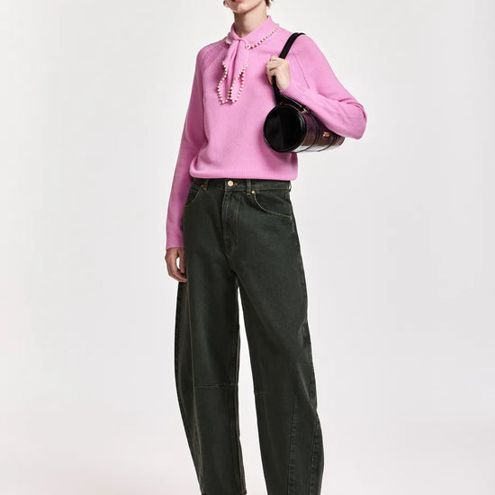 A model stands against a light gray studio background wearing a pink long-sleeve blouse with a tie-neck detail, dark olive green wide-leg jeans, black pointed shoes, and a black baseball cap, while carrying a black shoulder bag.