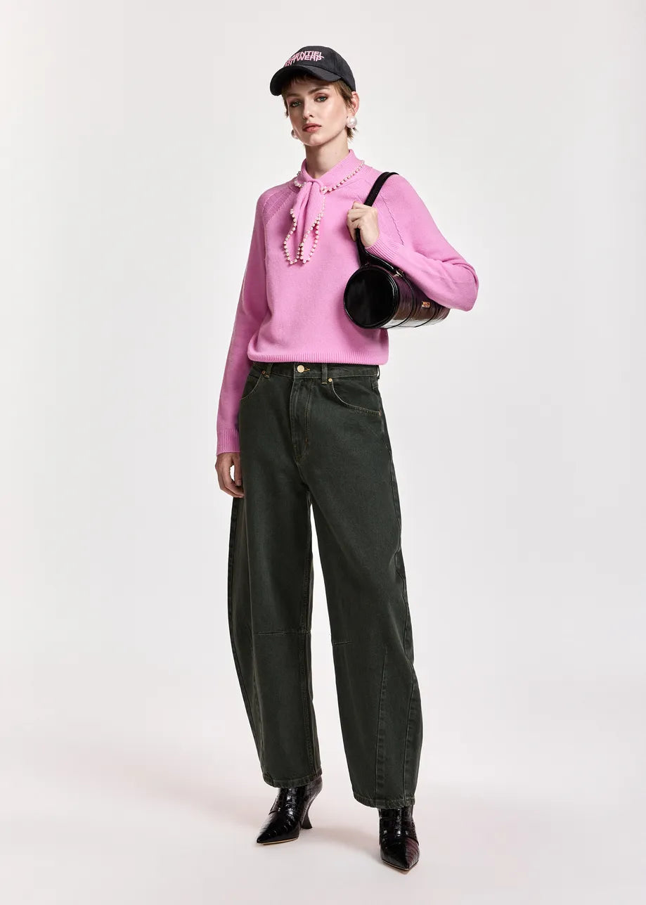A model stands against a light gray studio background wearing a pink long-sleeve blouse with a tie-neck detail, dark olive green wide-leg jeans, black pointed shoes, and a black baseball cap, while carrying a black shoulder bag.