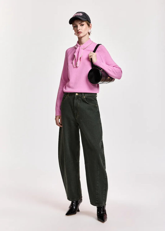 A model stands against a light gray studio background wearing a pink long-sleeve blouse with a tie-neck detail, dark olive green wide-leg jeans, black pointed shoes, and a black baseball cap, while carrying a black shoulder bag.