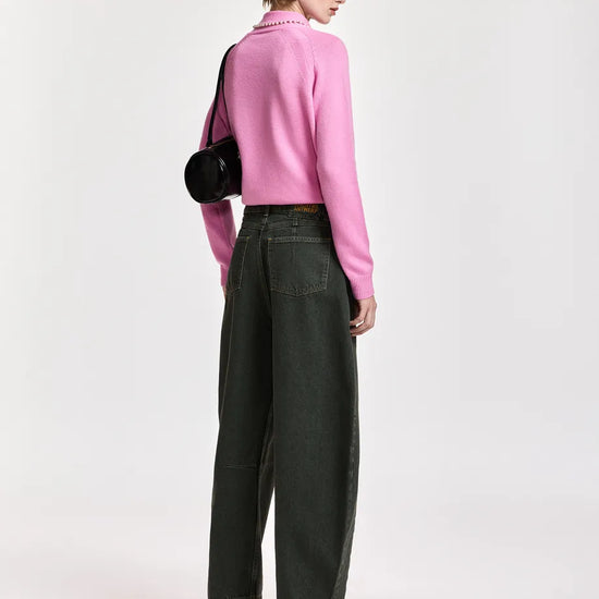 A model is shown from the back wearing a pink ribbed knit mock-neck sweater, dark olive green wide-leg jeans, black pointed-toe ankle boots, a black baseball cap, and carrying a black shoulder bag against a light gray studio background.