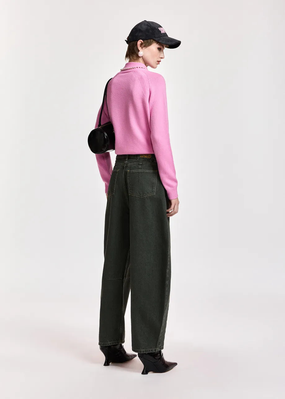 A model is shown from the back wearing a pink ribbed knit mock-neck sweater, dark olive green wide-leg jeans, black pointed-toe ankle boots, a black baseball cap, and carrying a black shoulder bag against a light gray studio background.