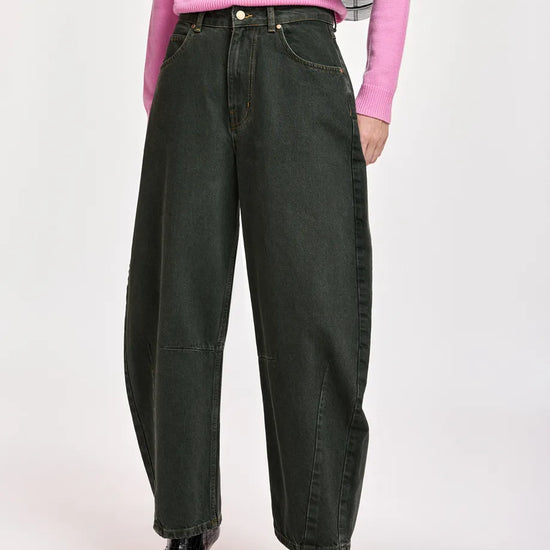 A model wearing dark olive green wide-leg denim jeans, styled with a pink sweater, black pointed-toe boots, and a black crossbody bag against a light gray studio background.