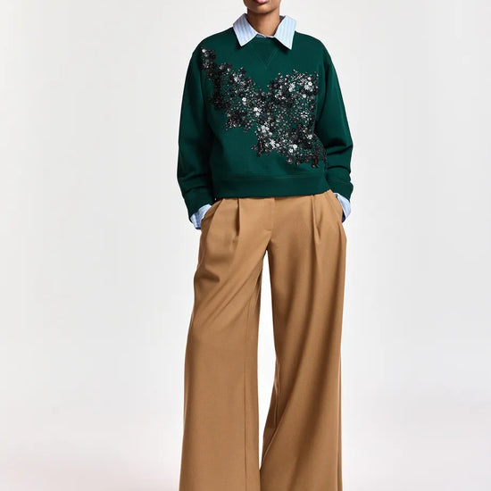 A model stands against a light gray studio background wearing a dark green crewneck sweater with a black and silver sequined butterfly motif across the chest, tucked into wide-leg tan trousers and paired with a light blue collared shirt, a patterned baseball cap, and black pointed shoes.