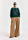 A model stands against a light gray studio background wearing a dark green crewneck sweater with a black and silver sequined butterfly motif across the chest, tucked into wide-leg tan trousers and paired with a light blue collared shirt, a patterned baseball cap, and black pointed shoes.
