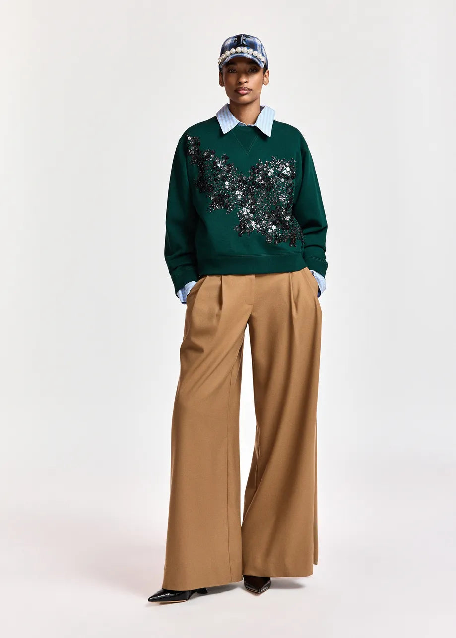 A model stands against a light gray studio background wearing a dark green crewneck sweater with a black and silver sequined butterfly motif across the chest, tucked into wide-leg tan trousers and paired with a light blue collared shirt, a patterned baseball cap, and black pointed shoes.
