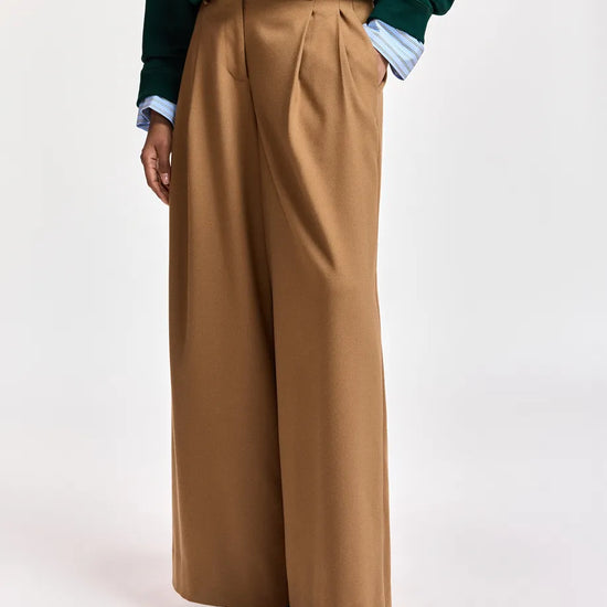 A person wearing high-waisted, wide-leg camel brown trousers, styled with a dark green sequined sweater and a light blue collared shirt, photographed against a plain white studio background.