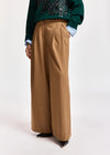A person wearing high-waisted, wide-leg camel brown trousers, styled with a dark green sequined sweater and a light blue collared shirt, photographed against a plain white studio background.