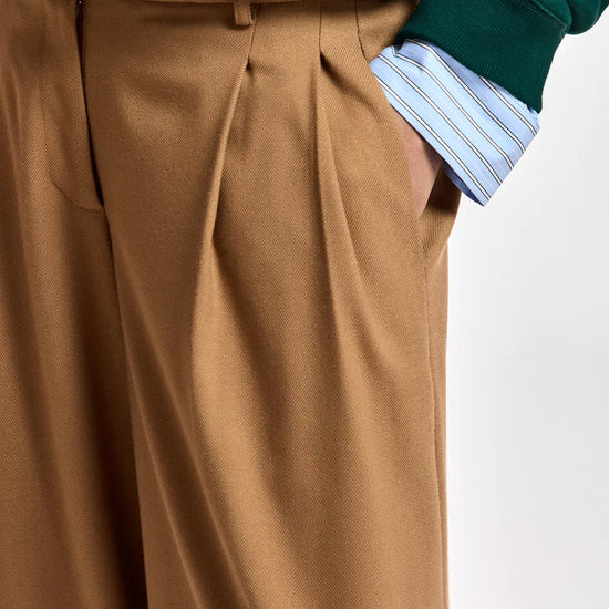Close-up of a person wearing tan wide-leg pleated trousers, a dark green sequined sweater, and a light blue and white striped button-down shirt, with one hand in the trouser pocket.