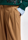 Close-up of a person wearing tan wide-leg pleated trousers, a dark green sequined sweater, and a light blue and white striped button-down shirt, with one hand in the trouser pocket.