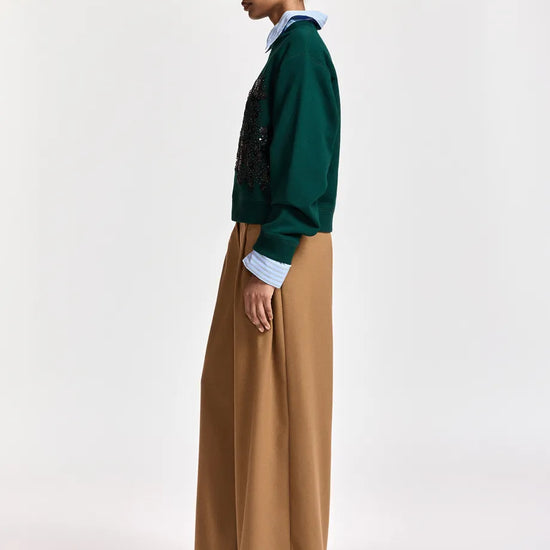A model shown in profile wearing a dark green knit cardigan with black sequined embroidery, a light blue collared shirt underneath, wide-leg tan trousers, and a blue and white plaid baseball cap, set against a plain light gray studio background.